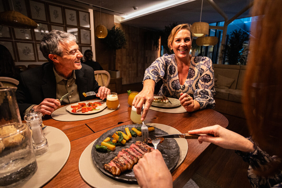 Paar sitzt in einem Restaurant in größerer Runde und genießt ein Steak. Neue TheFork-Daten zeigen, wie sich Restaurantbesuche auf Reisen verändern.